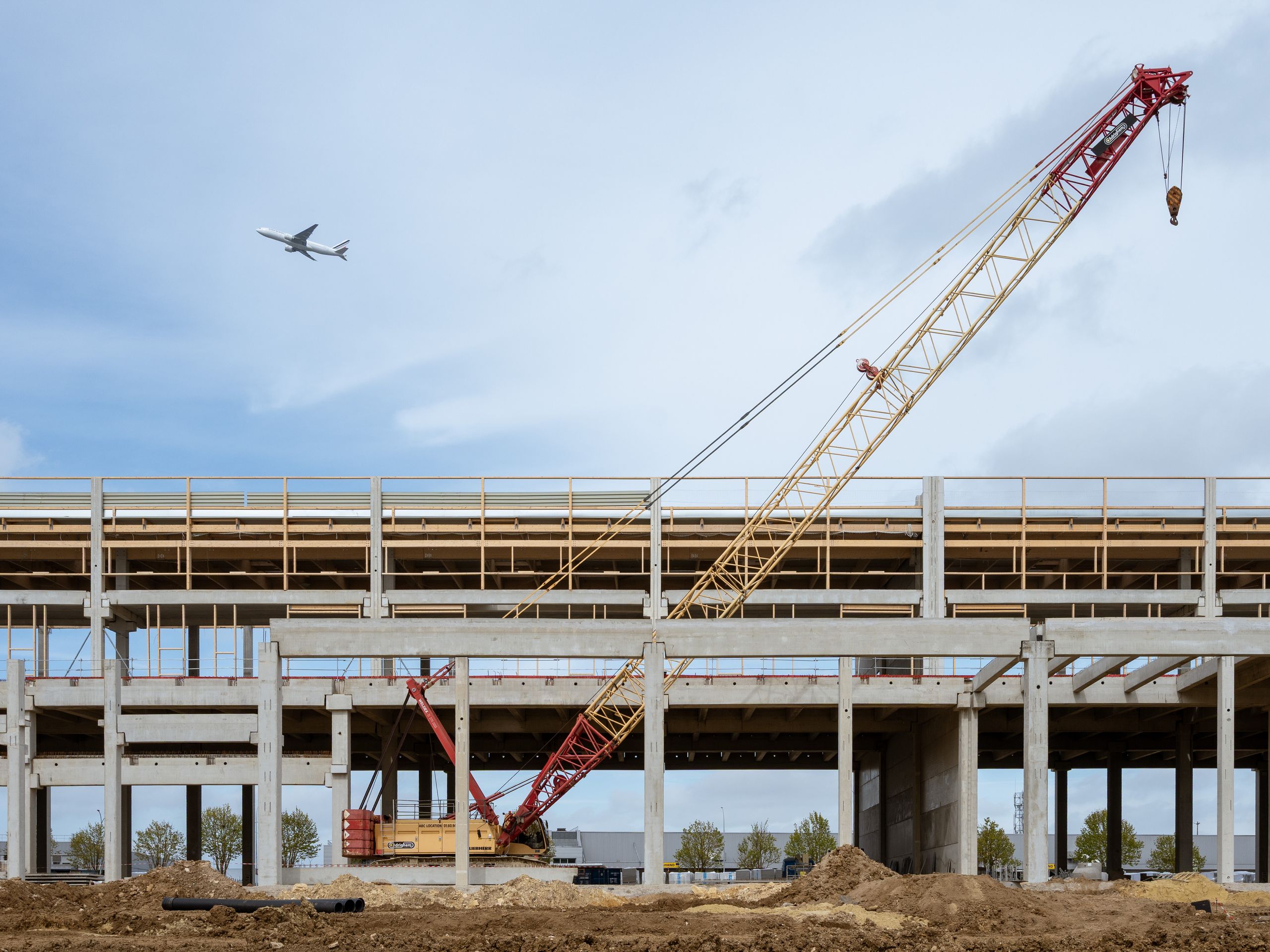 A large crane at a construction site with a plane flying overhead, set against a clear sky.